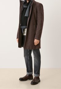 Cappotto di lana marrone scuro con una finitura testurizzata, indossato sopra un maglione nero e jeans blu. Accentuato da stivali in pelle marrone e una sciarpa grigia.