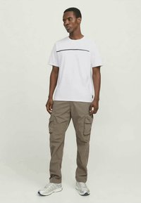 Homem vestindo uma T-shirt branca com uma faixa preta, calças cargo caqui e tênis cinza, de pé com as mãos relaxadas e olhando para o lado.