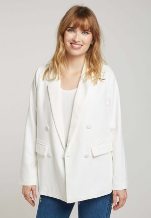 Blazer blanc à double boutonnage avec des revers crantés et des poches avant, présentant une texture douce et une coupe décontractée. Porté par-dessus un haut léger.