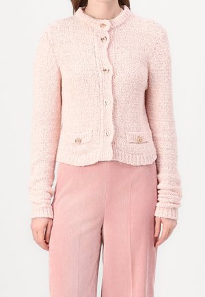 Vrouw draagt een lichtroze, gestructureerde, gebreide cardigan met knopen en bijpassende roze, geribbelde, wijd uitlopende broek, getoond van nek tot heupen.