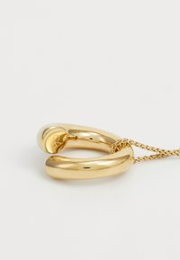Joyería de metal dorado que presenta dos formas circulares superpuestas con un acabado suave y brillante, y una cadena que une las dos piezas.