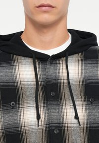 Ruudullinen flanellipaita musta-beige-väreillä, jossa on huppu, etunapit ja hienovarainen rakenne. Napit ovat mustat.