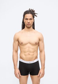 Άνδρας με ντραντλοκς όρθιος, φοράει μαύρα εσώρουχα Tommy Hilfiger, κοιτάζοντας μπροστά πάνω σε απλό ανοιχτό φόντο.