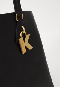 Geantă de mână din piele neagră, cu un etichetă metalică aurie ce prezintă un logo proeminent cu litera "K" și detalii texturate pe suprafață.