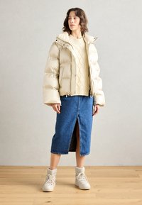 Jasno beżowa kurtka puchowa z kapturem, noszona na kremowym swetrze z dzianiny oraz dżinsowej spódnicy z przodem rozciętym. Całość dopełniają kremowe buty sznurowane.