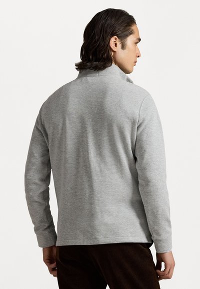 Άνδρας με μακριά σκούρα μαλλιά που φοράει ένα ανοιχτό γκρι πουλόβερ με μακριά μανίκια και σκούρες καφέ παντελόνες, γυρνώντας προς το μέρος ενός απλού λευκού φόντου.