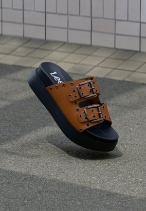 Καφέ δερμάτινο σανδάλι slide με δύο λουριά με μεταλλικές αγκράφες και παχύ μαύρο πέλμα σε πλακόστρωτη εξωτερική επιφάνεια.