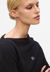 Schwarzer gestrickter Pullover mit hohem Kragen und einem gestickten weißen Logo auf der Brust. Weiche Textur, taillierte Silhouette und lange Ärmel.