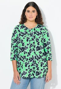 Zielona bluzka z dekoltem w serek i rękawami 3/4, ozdobiona czarno-białym abstrakcyjnym wzorem. Miękki materiał; luźny krój, połączona z jasnoniebieskimi dżinsami.