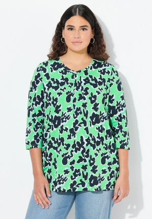 Haut vert à manches 3/4 avec col en V, présentant un motif abstrait noir et blanc. Tissu doux ; coupe décontractée, associé à un jean bleu clair.