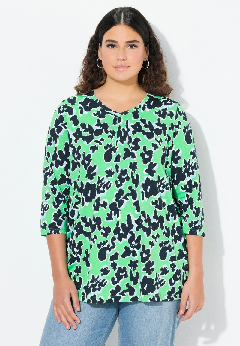 Zielona bluzka z dekoltem w serek i rękawami 3/4, ozdobiona czarno-białym abstrakcyjnym wzorem. Miękki materiał; luźny krój, połączona z jasnoniebieskimi dżinsami.