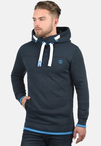 Donkerblauwe hoodie gemaakt van een katoenmix. Heeft een capuchon met trekkoord, blauwe accenten en een logo op de borst. Zachte textuur met een relaxte pasvorm.