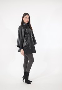 Czarna kurtka w stylu peleryny z sztucznej skóry, z wysokim kołnierzem, taliowana paskiem. Rozszerzane rękawy, gładka faktura, bez wzorów.