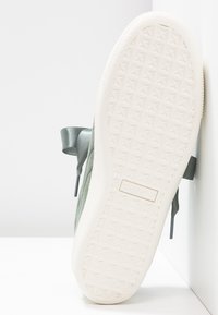 Weiße Sohle eines grünen Puma-Sneakers mit geometrischem Profil und satinierten Schnürsenkeln, schräg aufrecht vor einem weißen Wand- und Bodenhintergrund.