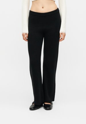 Vrouw draagt zwarte brede gebreide broek, witte cropped top met lange mouwen en zwarte platte schoenen met bandjes, staand tegen een effen achtergrond.