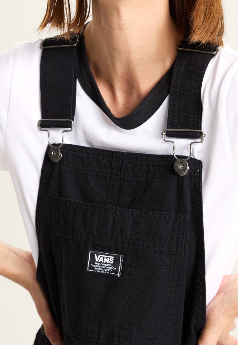 Personne portant une salopette Vans noire avec des boucles métalliques ajustables sur un t-shirt blanc, les mains posées sur les hanches, visible du cou à la taille.
