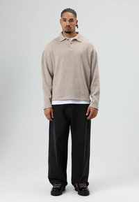 Pull beige en tricot avec col, manches longues et deux boutons ; porté sur un sous-vêtement blanc. Associé à un jean noir ample et des chaussures noires.