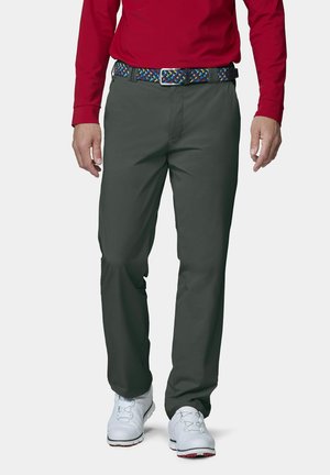Mann, der dunkelgraue Hose, bunten gewebten Gürtel, rotes langärmeliges Hemd und weiße Golfschuhe trägt, steht vor einem einfarbigen Hintergrund.