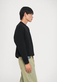 Felpa nera con maniche lunghe e scollo tondo, dotata di tasca frontale. Abbinata a pantaloni in denim chiaro e texturizzati di colore verde.