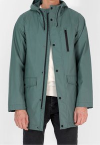 Mężczyzna w turkusowej kurtce przeciwdeszczowej z kapturem, z czarnym zamkiem i zatrzaskami, z kieszenią na zamek na przedniej piersi oraz kieszeniami na klapie, nosi białą bluzę i czarne spodnie.