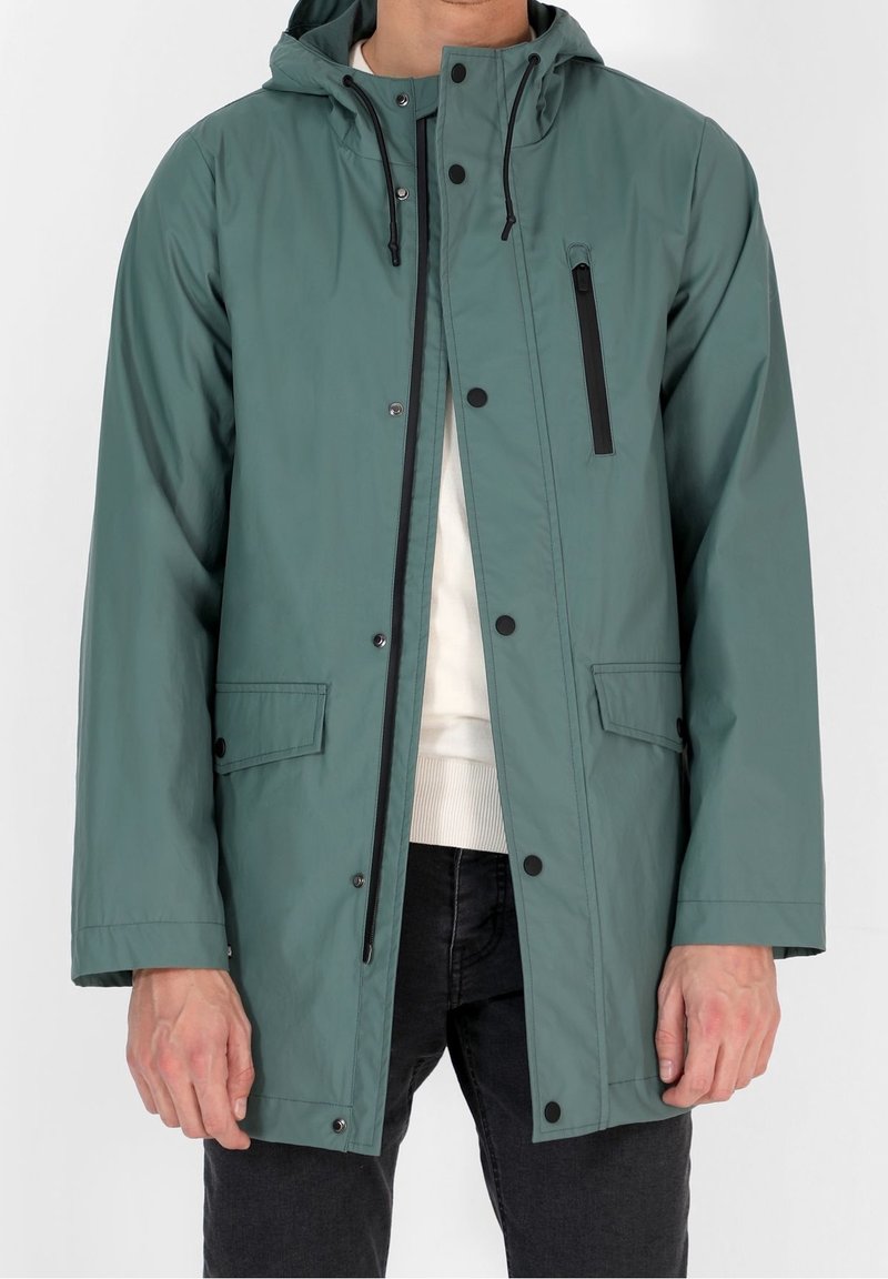 Mężczyzna w turkusowej kurtce przeciwdeszczowej z kapturem, z czarnym zamkiem i zatrzaskami, z kieszenią na zamek na przedniej piersi oraz kieszeniami na klapie, nosi białą bluzę i czarne spodnie.