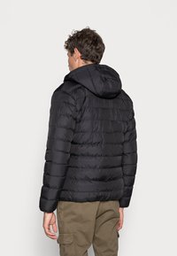 Black puffer jacket featuring a hood, quilted design with horizontal lines, and a smooth, shiny texture. Worn over khaki pants.