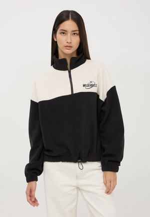 Pullover zip-up nero e beige realizzato in pile, con polsini elasticizzati, orlo con coulisse e un logo con il testo "WILDERNESS."