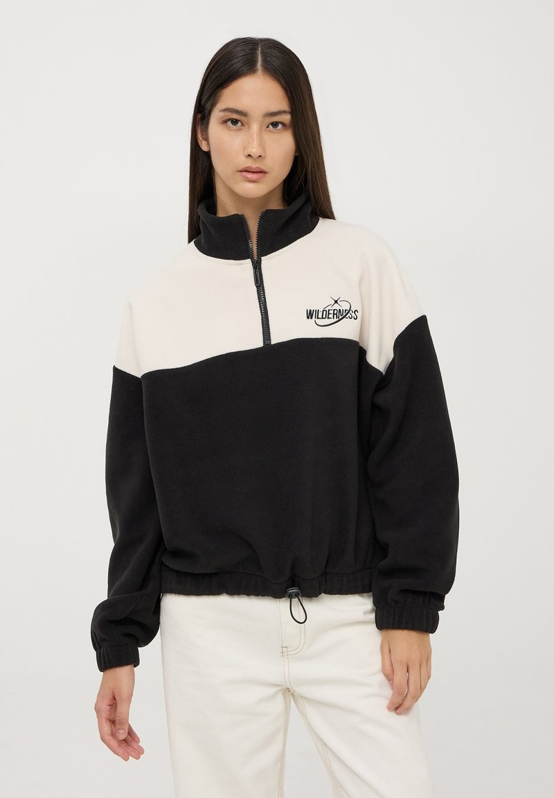 Pullover zip-up nero e beige realizzato in pile, con polsini elasticizzati, orlo con coulisse e un logo con il testo "WILDERNESS."
