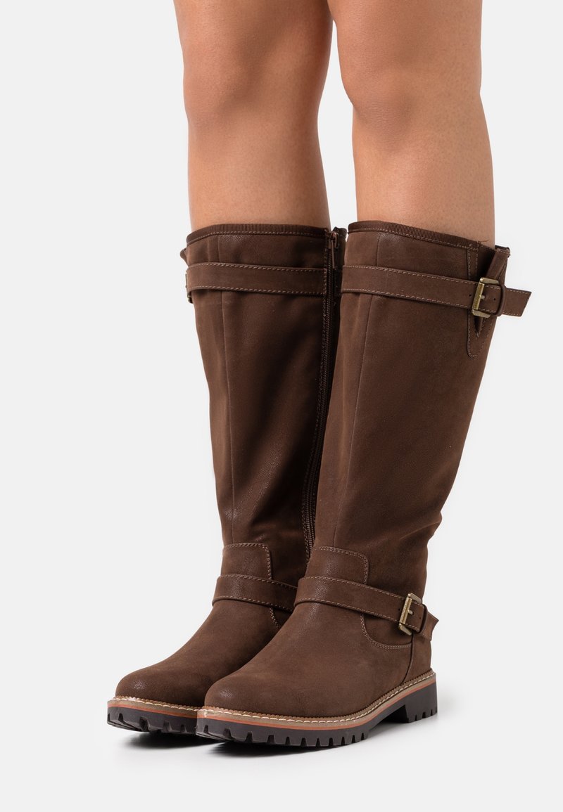 Bottes hautes en cuir marron avec des sangles bouclées à la cheville et près du haut, portées sur des jambes nues, posées sur un fond blanc.