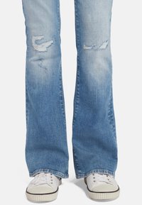 Jeans azzurri a zampa d'elefante con toppe visibili e una texture sbiadita, abbinati a sneakers bianche con suola a righe in gomma.
