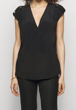 Kobieta ubrana w czarną, bezrękawną bluzkę z dekoltem w serek, z zapinanymi na guziki patkami na ramionach, oraz czarne dopasowane spodnie, stojąca na jednolitym tle.