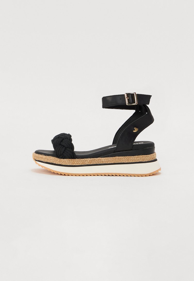 Black wedge sandals with a knotted rope accent, adjustable ankle strap, and a textured cork midsole. Contrasting white and gum rubber outsole.