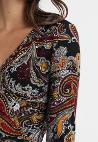 Primer plano de una prenda de manga larga con escote en V, con intrincados patrones de paisley en negro, burdeos, amarillo mostaza y blanco.