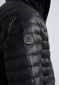 Veste matelassée noire avec un design matelassé, une texture brillante et un patch logo circulaire sur l'épaule avec des graphismes de montagne.