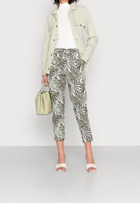 Světle zelená denimová bunda přes bílý top, kombinovaná s vzorovanými kalhotami s palmami v tmavě zelené a bílé, doplněná světle zelenou taškou.