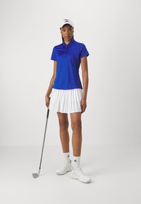 Modra polo majica, bela nabirana krila, bele golf čevlje in kapo. Stoji z golf palico, teksturirani materiali, športen kroj, sproščen dizajn.