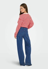 Dugi top s crvenim i bijelim prugama uparan s visokim strukom plavim trapericama širokih nogavica. Značajni detalji uključuju bočne džepove i šavove.