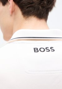 Λευκό πόλο πουκάμισο με υφασμάτινη υφή, με αντιθετικές μαύρες και καφέ ρίγες στον γιακά και κεντημένο λογότυπο "BOSS" στην πλάτη.