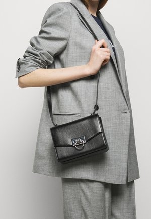 Osoba w szarłym garniturze trzyma czarną skórzaną torbę na ramię z srebrnym zapięciem w kształcie pierścienia, na jednolitym jasnym tle.