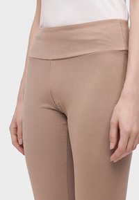 Gros plan d'une personne portant des leggings beige taille haute et un haut blanc, les mains détendues le long du corps.