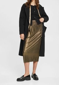 Zwarte jas over een zwarte top, gecombineerd met een metallic gouden geplooide rok en zwarte lakleren schoenen. Vloeiende stof, midi lengte.