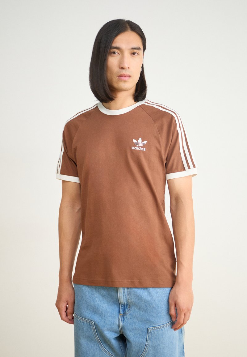 Ruskea puuvillapaita, jossa on valkoinen ribbineck, lyhyet hihat ja kolme valkoista raitaa olkapäillä. Paitaa koristaa pieni valkoinen Adidas-logo.