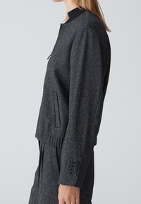 Femme portant un blazer gris foncé boutonné et un pantalon plissé, debout de profil devant un fond uni gris clair.