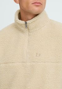 Beige fleece pullover med høj krave, halv lynlås fortil og en struktureret overflade. Har et diskret logo på brystet og kontrasterende sømme.