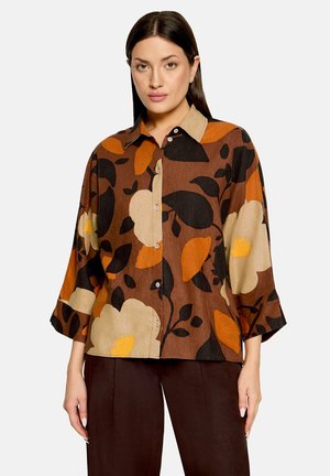 Femme aux cheveux bruns lisses portant une chemise marron à boutons avec de grands motifs floraux beige, orange et noirs et un pantalon marron foncé.