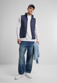 Marinefarbener, gesteppter Weste über einem weißen Langarmshirt, kombiniert mit lässigen blauen Jeans-Cargohosen und weißen Sneakers. Ein blauer Pullover ist über den Arm gelegt.