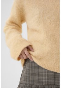 Weicher gelber gestrickter Pullover mit langen, ausladenden Ärmeln, kombiniert mit einem karierten Rock, der horizontale und vertikale Linien aufweist.