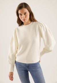 Felpa color crema con maniche a palloncino, scollo rotondo e polsini a coste. Abbinata a jeans in denim blu. Tessuto morbido e strutturato.