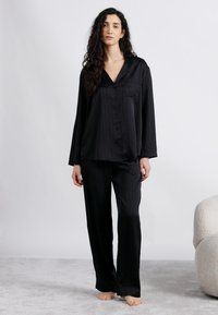 Γυναίκα που στέκεται ξυπόλυτη σε γκρι χαλί, φορώντας χαλαρή μαύρη σατέν πιτζάμα με μακριά μανίκια και παντελόνι, πάνω σε λευκό φόντο.