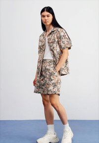 Camicia a maniche corte con bottoni e pantaloni corti abbinati in beige, con motivi astratti arancioni e neri. Il modello indossa una t-shirt bianca e sneakers.